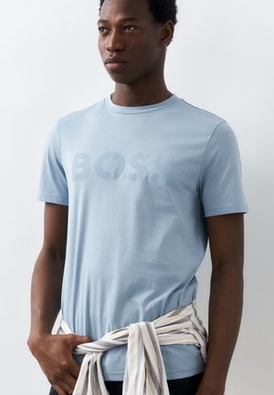 Άνδρας που φοράει ανοιχτό μπλε κοντομάνικο πουκάμισο με το κείμενο "BOSS", ριγέ πουκάμισο μακρυμάνικο δεμένο γύρω από τη μέση, στέκεται μπροστά σε λευκό φόντο.