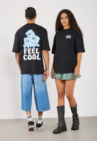 Schwarze übergroße T-Shirts mit cartoonhaften Eiswürfeln und dem Text "FEEL COOL". Kombiniert mit hellblauen Denim-Hosen und einem Denim-Rock. Dunkle Stiefel.