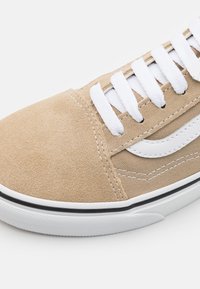 Sneaker in suede beige con lacci bianchi, striscia laterale bianca a contrasto e suola in gomma nera con un motivo testurizzato.