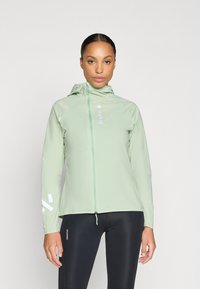 Chaqueta ligera de color verde claro con capucha, cierre completo, detalles reflectantes en las mangas y una textura suave. Combinada con mallas negras.