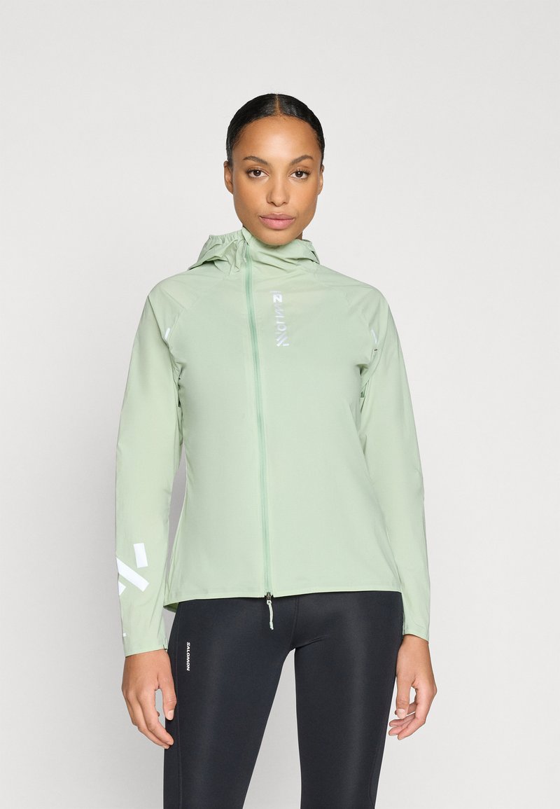 Chaqueta ligera de color verde claro con capucha, cierre completo, detalles reflectantes en las mangas y una textura suave. Combinada con mallas negras.