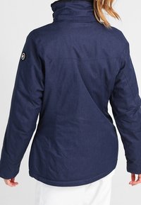 Marineblaue isolierte Jacke mit hohem Kragen, schwarzer Fütterung, Reißverschluss und einem Logopatch auf der linken Schulter.