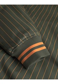 Grüner Stoff mit vertikalen orangefarbenen Streifen. Umschlagärmel mit geripptem Muster und orangen Akzenten. Glattes Material mit polierter Oberfläche.