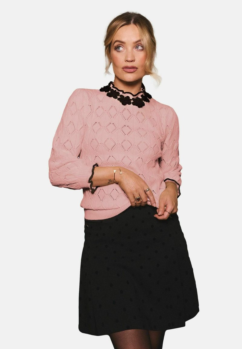 Pulover roz tricotat cu modele în formă de diamant și mâneci trei-sferturi, asortat cu o fustă neagră cu buline. Accesoriu cu guler floral negru.