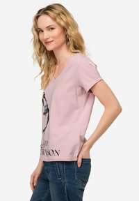Camiseta de cuello en V rosa hecha de una tela suave, con un estampado gráfico en el frente. Combinada con jeans de mezclilla azul, vista lateral del modelo.