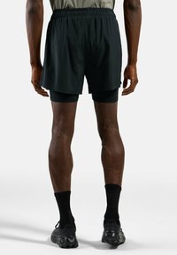 Zwarte sportshorts met een elastische tailleband, gedragen over zwarte aangesloten compressieshorts. Vergezeld van zwarte sportsokken en sneakers.