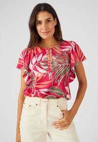 Top floral con patrones de hojas rojas y rosas, mangas cortas con volantes, cuello redondo y una tapeta con botones. Combinado con jeans de color claro.
