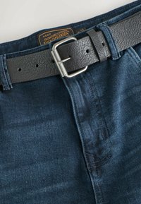 Tumed sinised teksapüksid musta nahkrihmaga, millel on hõbedane pandla. Nii pükstel kui rihmal on "Next Signature Denim Goods" silt vöökohtades.