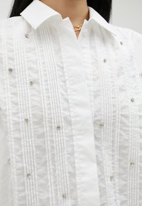 Chemise blanche avec un col structuré, présentant des plis verticaux et de petites accents métalliques décoratifs uniformément répartis sur le tissu.