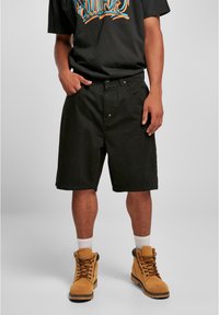 Sorte bomuldsshorts med to forlommer, afslappet pasform og knaplukning, parret med brune snørebånd støvler og hvide ankelsokker.