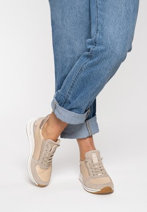 Person trägt beige Turnschuhe mit weißen Sohlen und hochgekrempelte blaue Jeans, schlägt ein Bein über das andere vor einem schlichten Hintergrund.