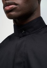 Camisa preta com colarinho alto, apresentando um pequeno fecho de botão e um tecido liso e suave. Close-up do detalhe do colarinho.