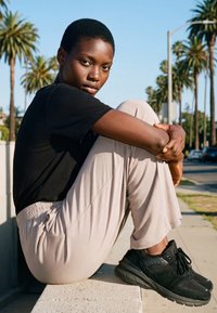 Ung person med kort hår iført sort skjorte og beige bukser sidder på en kant udendørs med palmer og blå himmel i baggrunden.