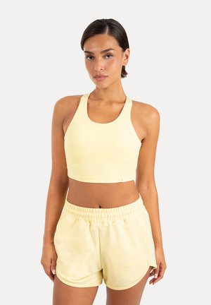 Kobieta stojąca, ubrana w bladożółty bezrękawnik typu crop top i pasujące krótkie spodenki z elastycznym pasem na jednolitym jasnym tle.