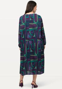 Pitkä, poimutettu mekko purppuran, vihreän ja sinisen sävyissä, jossa on abstrakti geometrinen kuvio. Mekossa on pitkät hihat ja venepäänteinen kaula-aukko.