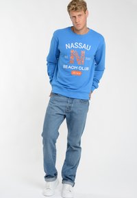 Niebieska bluza z nadrukiem "NASSAU BEACH CLUB Ibiza" w pomarańczowym i białym kolorze, zestawiona z jasnoniebieskimi dżinsami i białymi trampkami. Prosty design.