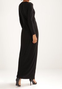 Kvinna iklädd en långärmad, golvlång svart klänning och svarta högklackade skor, stående mot en enkel ljus bakgrund.