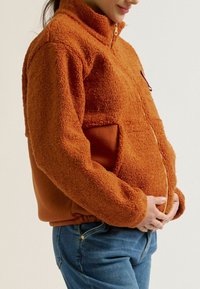 Oranger Fleece-Jacke mit hohem Kragen, Reißverschluss und seitlichen Taschen. Weiche, strukturierte Oberfläche. Elastischer Bund und kontrastierende Stoffdetails.