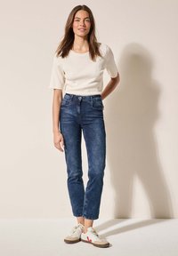 Haut léger crème à manches courtes, jean taille haute bleu foncé avec une coupe ajustée, baskets blanches avec des accents beige et rouge, fond neutre.