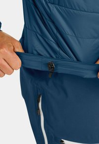 Dunkelblaue isolierte Jacke mit einer Reißverschlusstasche, die eine gesteppten Textur und eine Kordelzugverstellung am Saum zeigt.