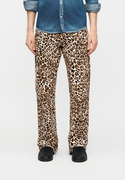 Άνδρας που φοράει παντελόνι με λεοπάρ σχέδιο, μαύρα παπούτσια και μπλε τζιν πουκάμισο με ρυμουλκιασμένα μανίκια, στέκεται μπροστά σε ένα απλό φόντο.