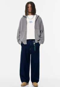 Grauer Strickzipper über einem weißen Langarmshirt, kombiniert mit weit geschnittenen dunkelblauen Jeans. Tanfarbene Stiefel und ein Accessoire mit grünen Akzenten.