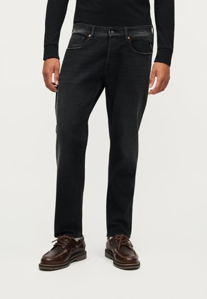 Homme portant un jean noir, un haut à manches longues noir et des chaussures en cuir marron à lacets, debout devant un fond blanc.