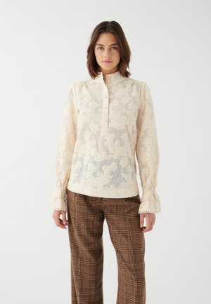 Blusa in pizzo color crema, maniche lunghe trasparenti con polsini arricciati, colletto alto con bottoni, motivo floreale su tutto il capo, abbinata a pantaloni a quadri marroni.