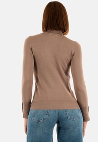Frau mit glattem braunem Haar, die ein enges taubfarbenes Langarmoberteil und blaue Jeans trägt, von hinten vor einem weißen Hintergrund gesehen.