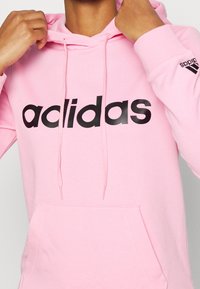 Różowa bluza z kapturem wykonana z mieszanki bawełny, z dużym czarnym logo "adidas" z przodu i żeberkowymi mankietami. Zawiera sznurki oraz kieszeń kangurzą.