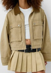 Khaki krótka kurtka z sztruksowym kołnierzem, dwiema przednimi kieszeniami i srebrnymi detalami, w połączeniu z plisowaną beżową spódnicą. Teksturowany materiał.