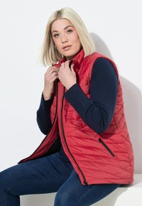 Rode gewatteerde vest met hoge kraag, rits aan de voorkant en twee ritszakken. Draagt over een marineblauw shirt met lange mouwen, met een gewatteerde textuur.
