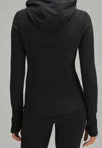 Žena v upnuté černé bundě s kapucí a otvory na palce, v černých legínách, zobrazená zezadu na jednobarevném pozadí.