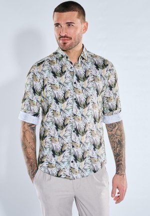 Mann mit kurzen Haaren und Tattoos, der ein kurzärmliges, geknöpftes Hemd mit tropischem Blattmuster und hellfarbene Hosen trägt, stehend mit den Händen in den Taschen.