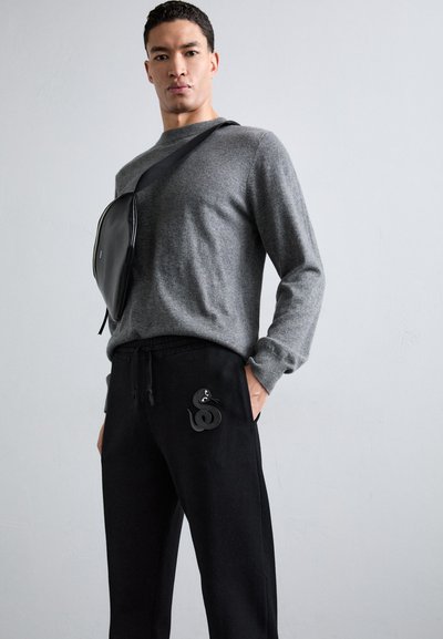 Grauer Pullover, schwarze Jogginghose mit Schlangenpatch und eine schwarze Umhängetasche. Das Material scheint weich und der Schnitt ist lässig.