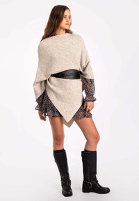 Poncho de punto beige que cae sobre un vestido floral oscuro, ceñido con un cinturón de cuero negro con tachuelas. La modelo lleva botas negras hasta la rodilla.