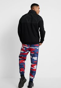 Giacca nera con zip e pannello lucido sul retro; abbinata a pantaloni tuta blu a motivo camouflage con dettagli rossi e bianchi; sneakers nere.