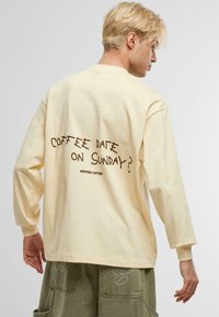 T-shirt en coton à manches longues couleur crème avec un graphisme textuel ludique de couleur marron au dos qui dit "Rendez-vous café le dimanche ?"