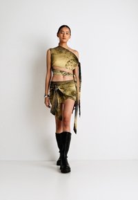 Zeleno-hnedý vzorovaný crop top s asymetrickým dizajnom, skombinovaný s rovnako ladenou zavinovacou sukňou a čiernymi vysokými čižmami po kolená.