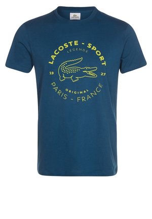T-shirt bleu Lacoste Sport avec logo crocodile jaune et texte indiquant "Lacoste - Sport," "Legende," "Original," et "Paris - France 1927."