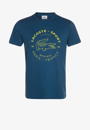 T-shirt bleu Lacoste Sport avec logo crocodile jaune et texte indiquant "Lacoste - Sport," "Legende," "Original," et "Paris - France 1927."