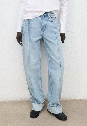 Una persona indossa jeans a gamba larga di un blu chiaro con risvolti sopra scarpe nere e una maglietta bianca a maniche lunghe infilata, mentre si trova in piedi contro un muro bianco.