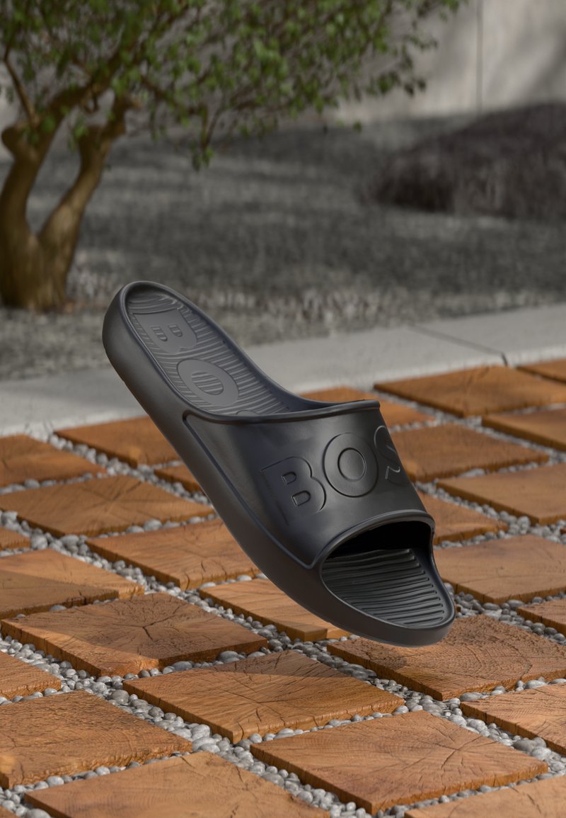 Chinelo de borracha preto com biqueira aberta, palmilha texturizada e logótipo "BOS" estampado. Apresentado sobre um fundo de azulejos de madeira e cascalho.