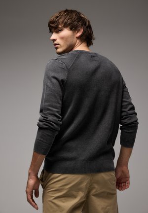 Grauer Strickpullover mit gerippter Textur, Rundhalsausschnitt und langen Ärmeln, getragen über khakifarbenen Hosen. Verfügt über eine taillierte Silhouette und subtile Nahtdetails.