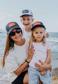 Three people pose by the water. The adult wears a light shirt and sunglasses. Two children wear colorful caps and casual clothing.