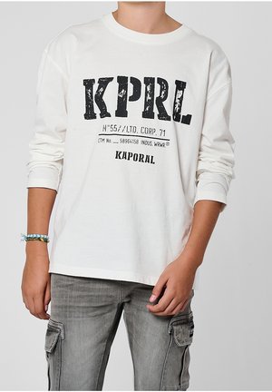 Person iført en hvid langærmet skjorte med sort "KPRL" tekst og grå cargobukser, mod en ensartet lysegrå baggrund.