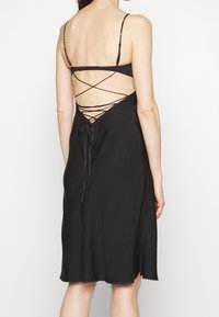Černé šaty s nastavitelnými špagetovými ramínky, hlubokým výstřihem na zádech a šněrováním s mašličkami. Tkanina vypadá hladce a lehce.