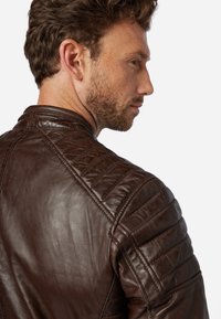 Brun läderjacka med en kviltad design på axlarna och en slät yta. Kragen är rundad med en stilren silhuett.