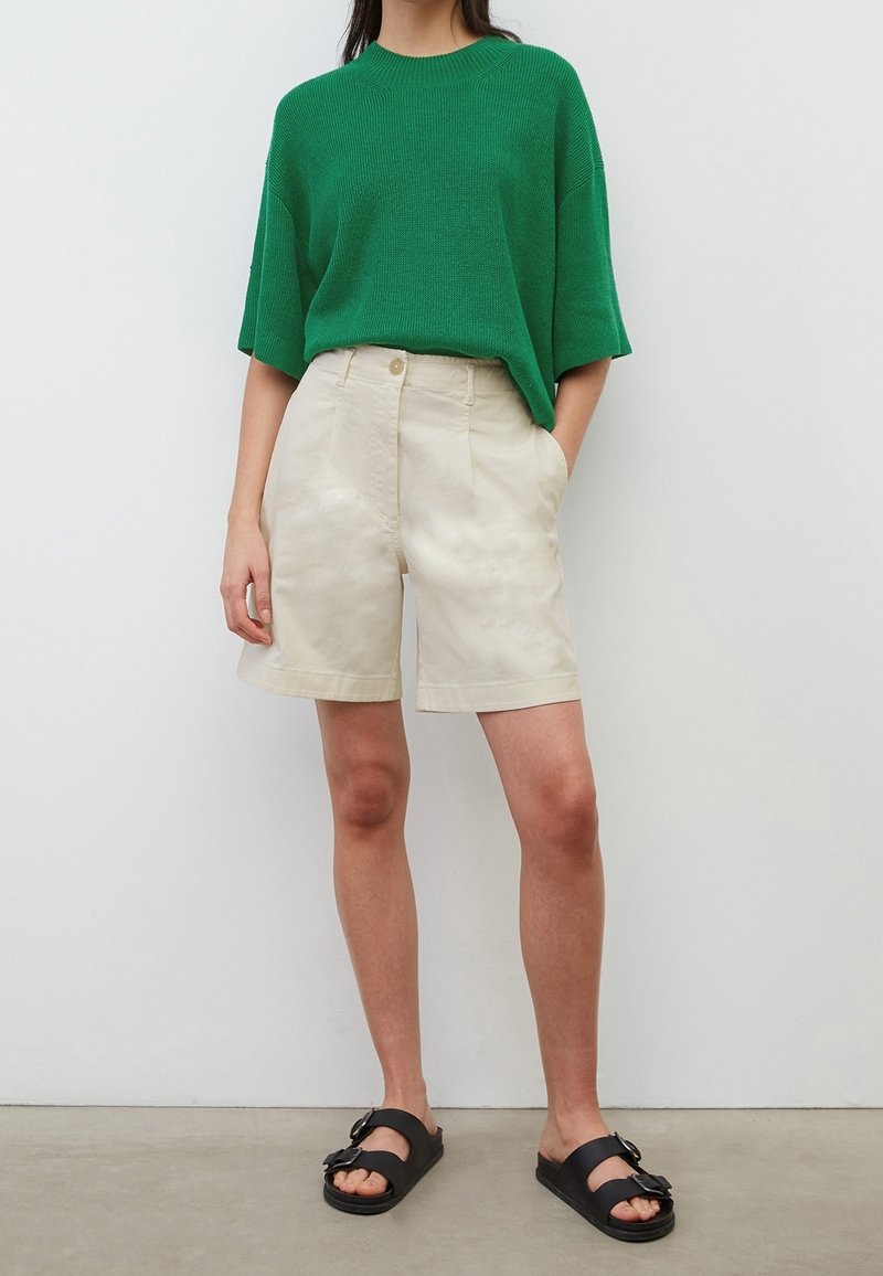 Personne portant un pull vert à manches courtes, un short beige jusqu'aux genoux, des sandales noires à boucle, debout contre un mur blanc uni.