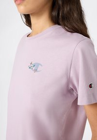 Kobieta ubrana w jasnofioletowy T-shirt z małym niebieskim logo przedstawiającym czaszkę z skrzydłami i różowym sercem na piersi oraz naszywką logo na rękawie.